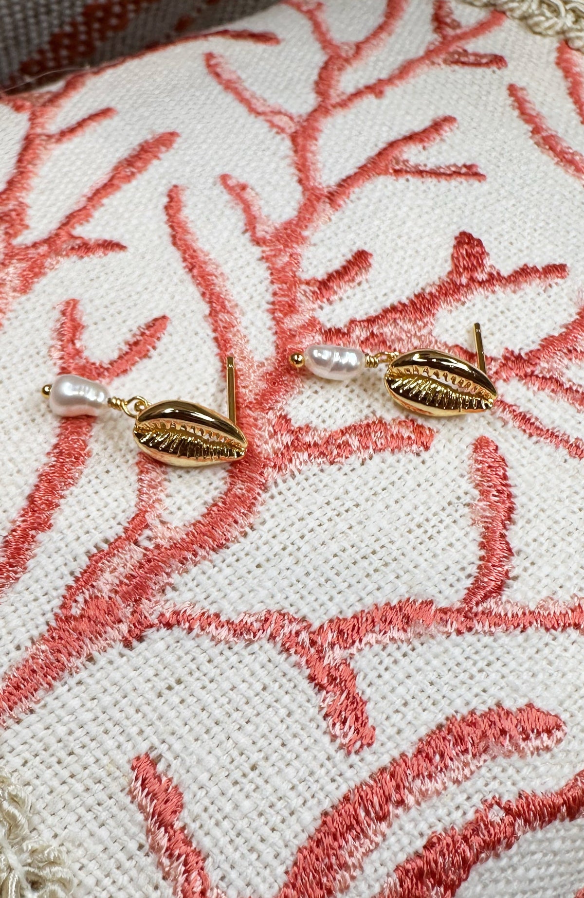 A pair of gold-toned leaf-shaped earrings with small white pearl drops, displayed on a textured cream fabric cushion with coral-pink embroidery.