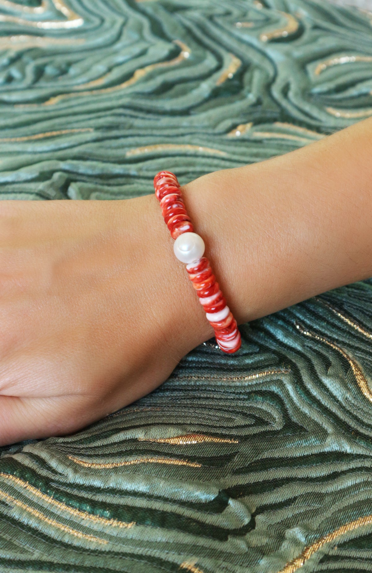 A close-up of a person's wrist wearing a beaded bracelet with red and white marbled beads and a single white pearl-like accent bead, set against a green patterned fabric background.