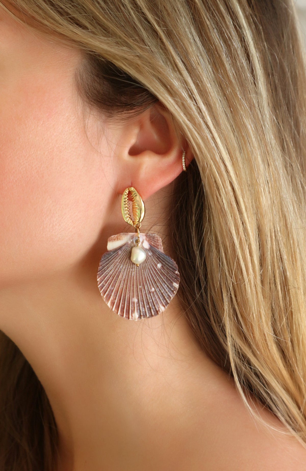 A close-up of a woman's ear wearing a dangling shell earring. The earring features a gold-toned top loop, a small pearl accent, and a larger pink shell charm with a pearl bead at the center.
