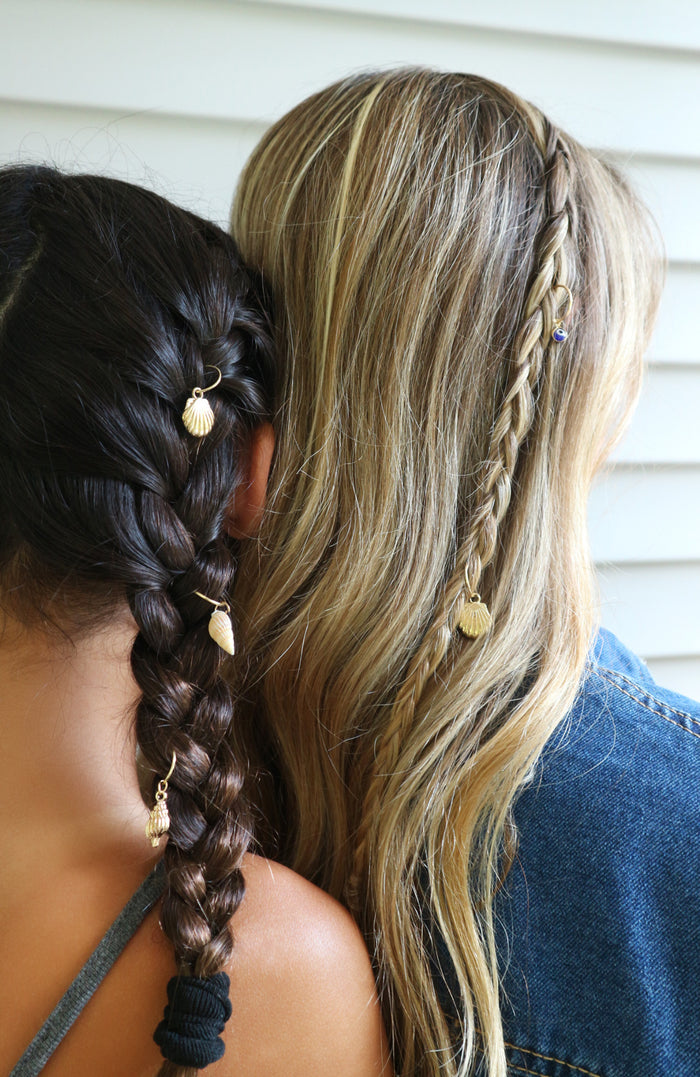 Two women shown from behind with detailed braids, one with dark brown hair and one with blonde hair, both styled with small gold shell and coin hair charms against a white siding background.