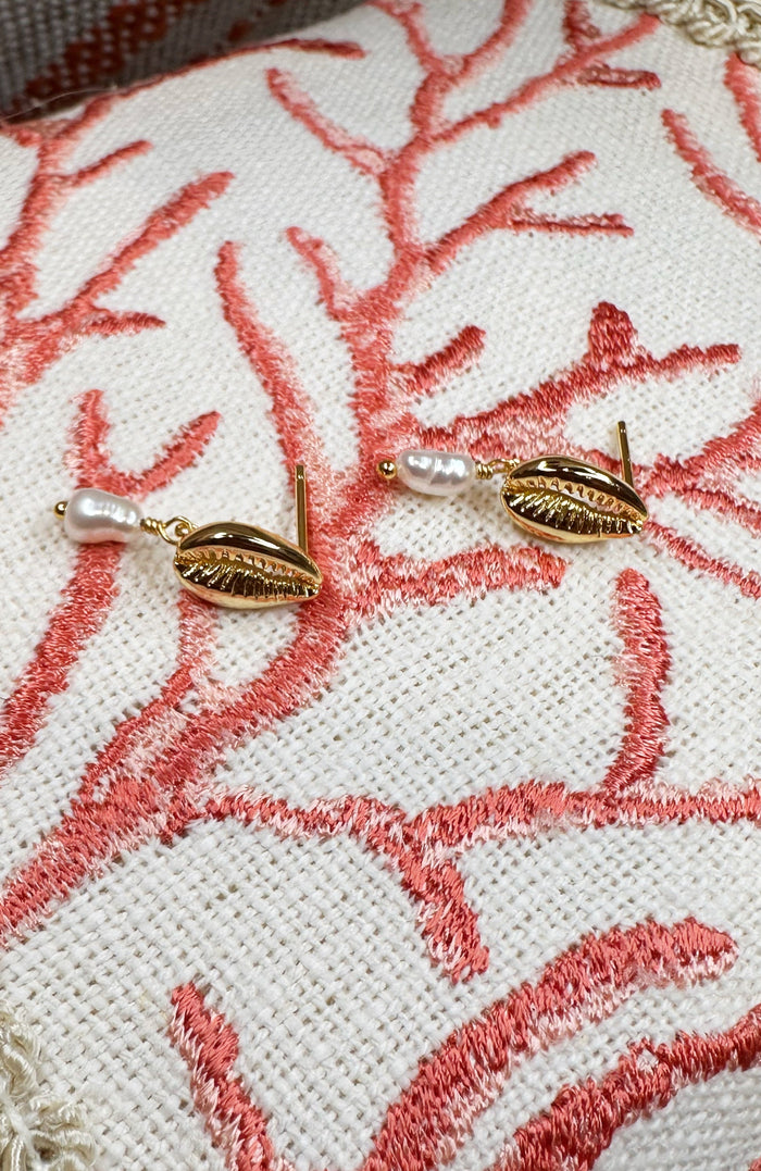 A pair of gold-toned leaf-shaped earrings with small white pearl drops, displayed on a textured cream fabric cushion with coral-pink embroidery.