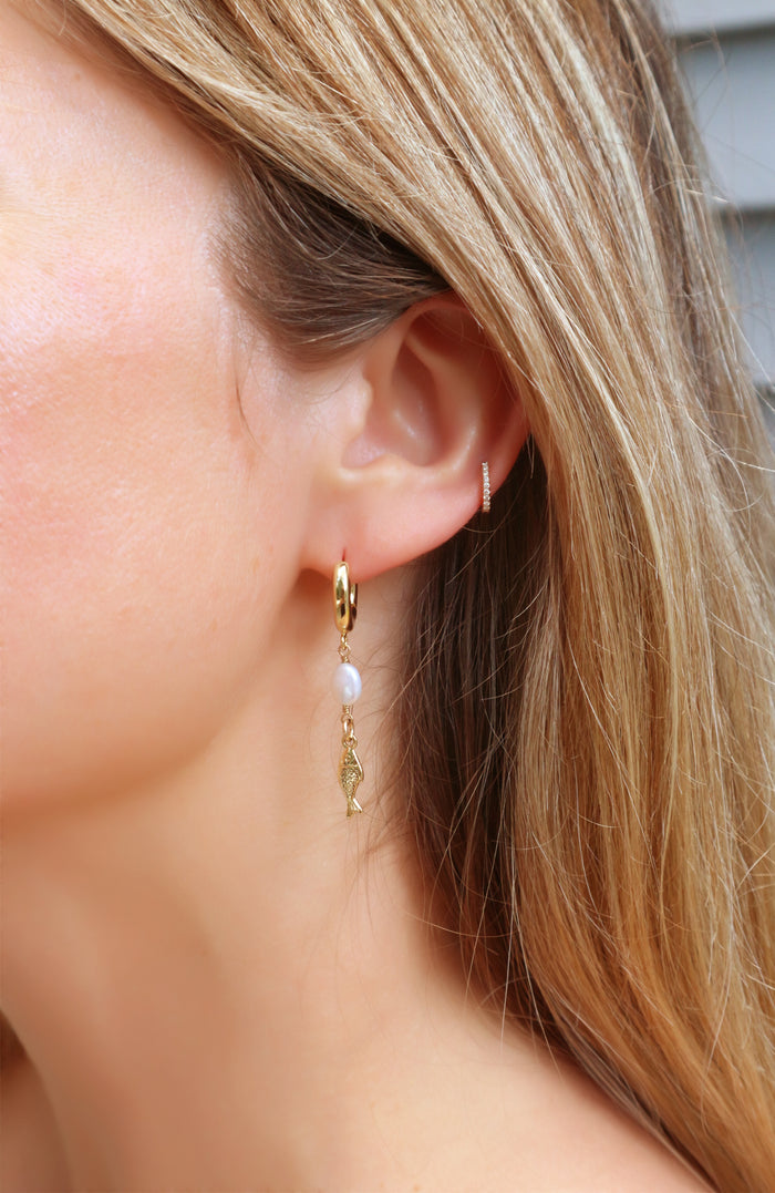 A close-up of a woman's ear wearing a delicate gold huggie hoop earring with a dangling white pearl and a small gold fish charm, alongside a tiny gold stud in her upper ear cartilage.