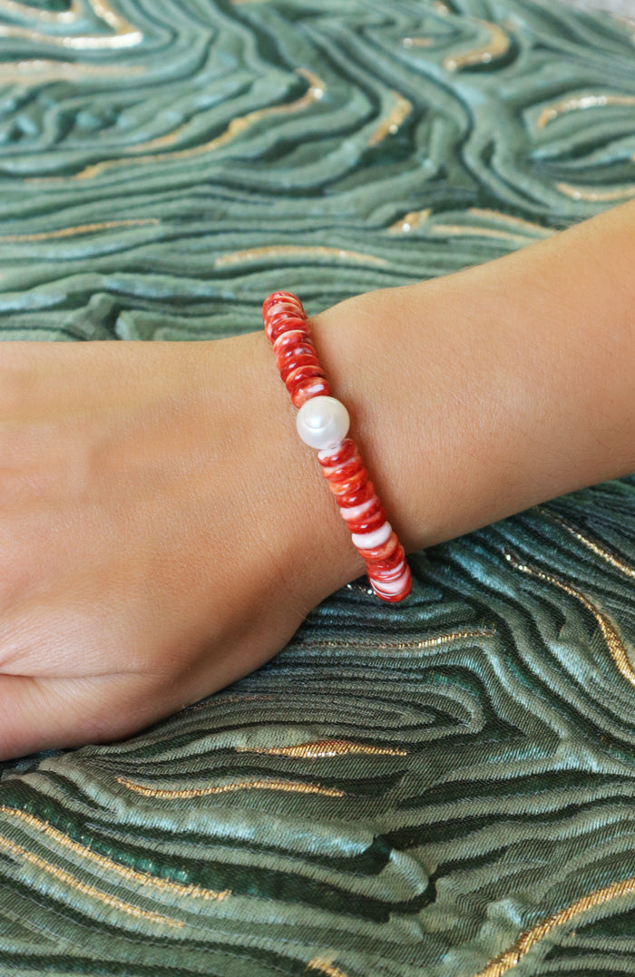 A close-up of a person's wrist wearing a beaded bracelet with red and white marbled beads and a single white pearl-like accent bead, set against a green patterned fabric background.