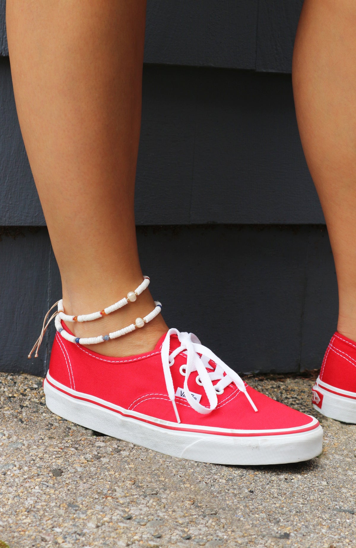 A close-up of a person's foot wearing a red canvas low-top sneaker with white laces and a white rubber sole, standing on a gravel surface in front of a dark gray wall. A beaded ankle bracelet is also visible.