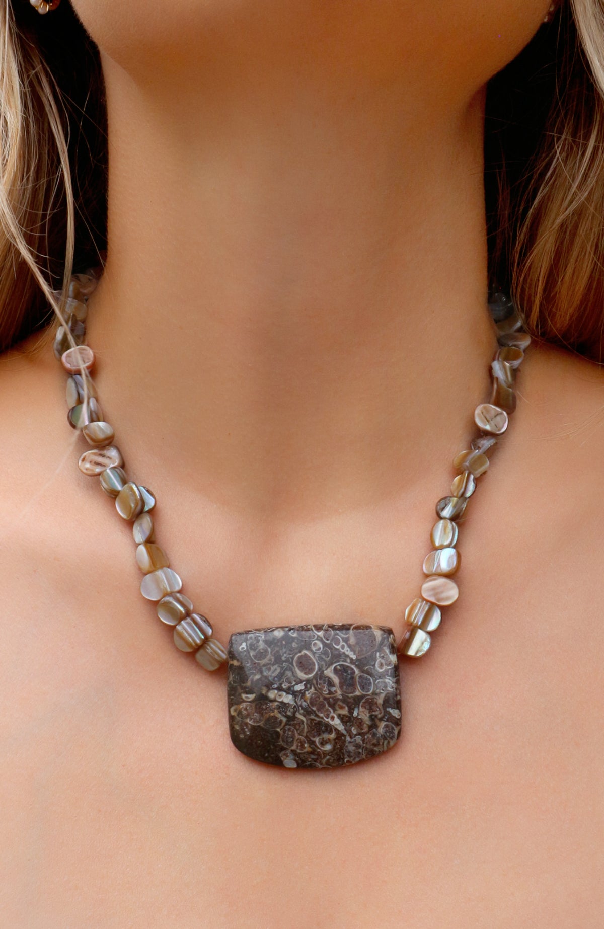 A close-up of a woman's neck and chest wearing a necklace with a large rectangular dark stone pendant and small iridescent shell-like beads along the chain.