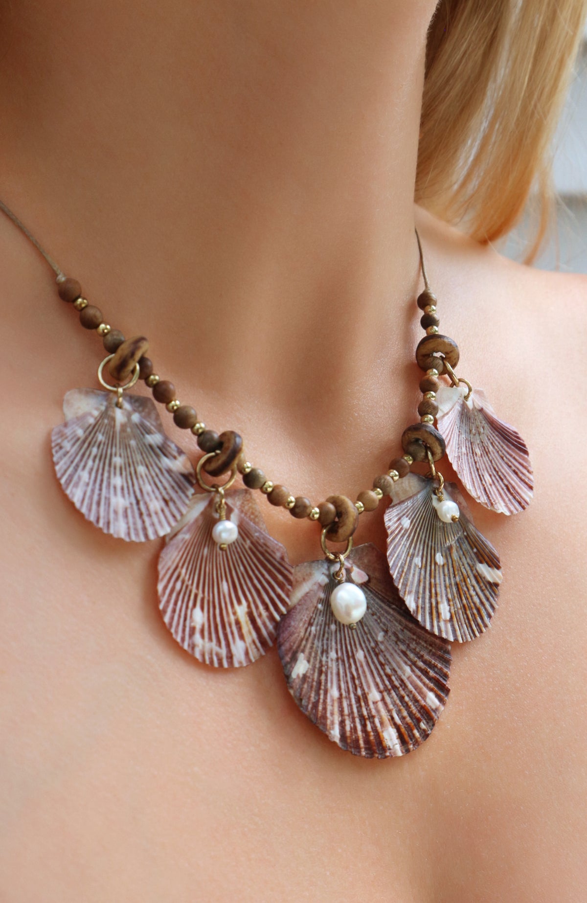 A close-up of a woman's neck and upper chest wearing a bohemian-style shell necklace. The necklace features five large, fan-shaped seashells in muted pink tones, accented with small brown beads and white pearl drops.