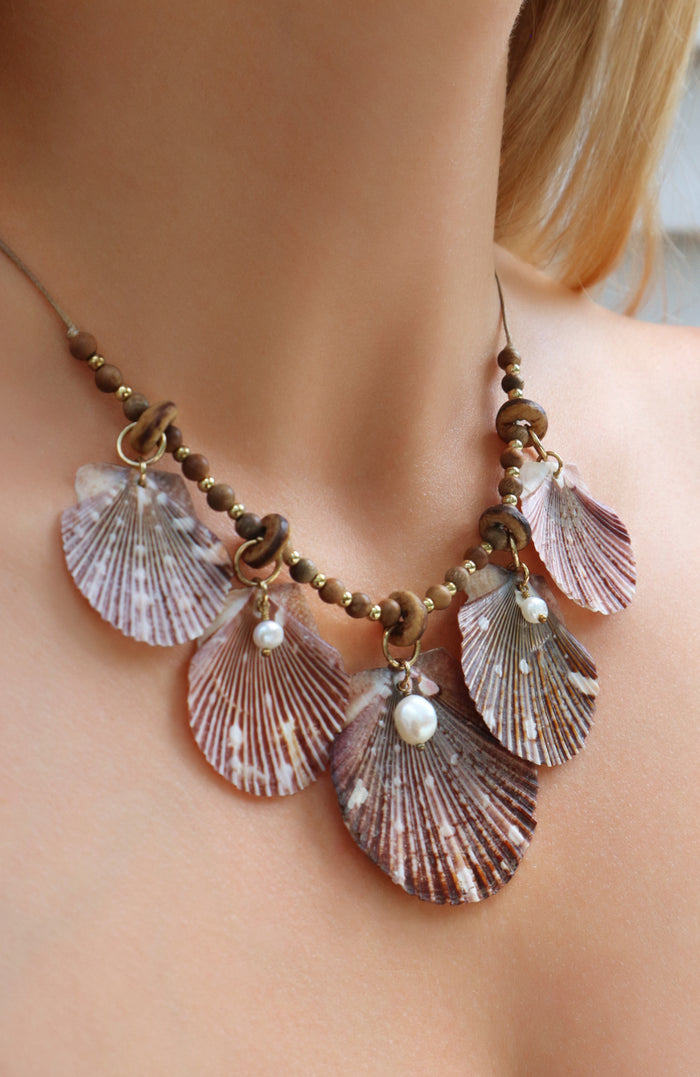 A close-up of a woman's neck and upper chest wearing a bohemian-style shell necklace. The necklace features five large, fan-shaped seashells in muted pink tones, accented with small brown beads and white pearl drops.