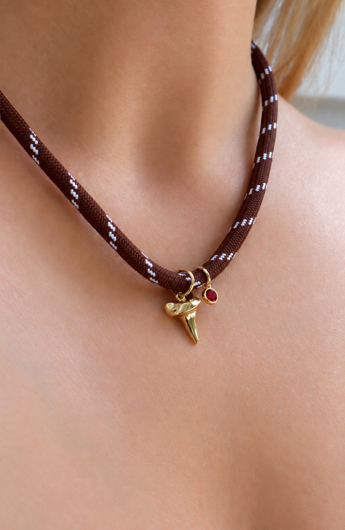 A close-up of a person's neck and chest wearing a brown braided cord choker necklace with a gold shark tooth charm and a small red gem accent.