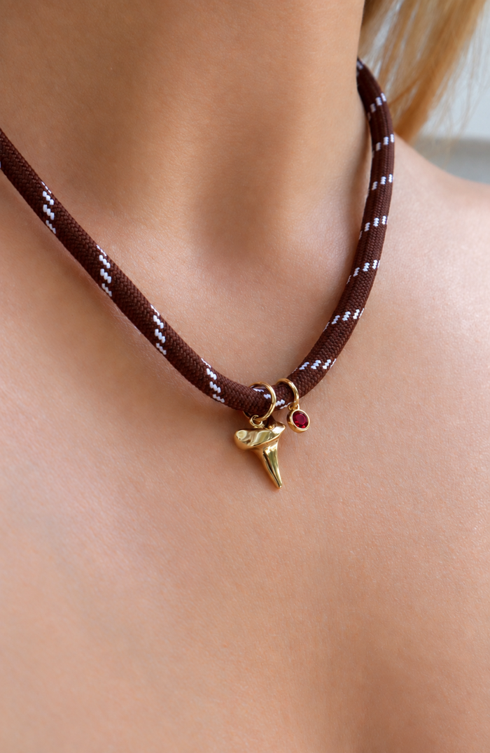 A close-up of a person's neck and chest wearing a brown braided cord choker necklace with a gold shark tooth charm and a small red gem accent.