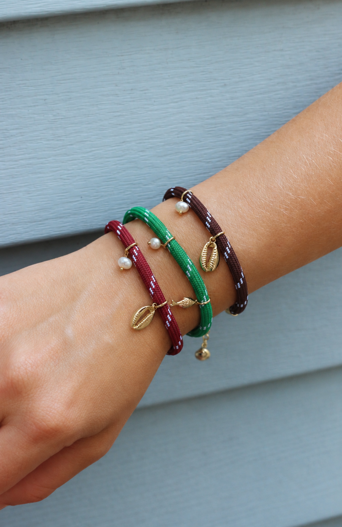 Three woven cord bracelets in burgundy, green, and dark brown, each decorated with small white beads and gold-toned leaf charms, shown worn on a person's wrist against a light blue-gray siding background.