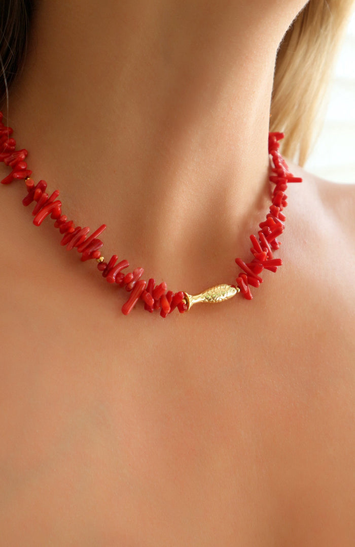 A close-up of a model's neck and upper chest wearing a red coral chip necklace with a small gold fish-shaped charm at the center.