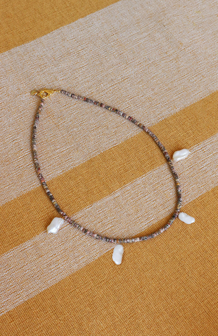 A beaded necklace with small multicolored beads and five white shell-like charms, displayed on a striped beige and mustard yellow fabric background.