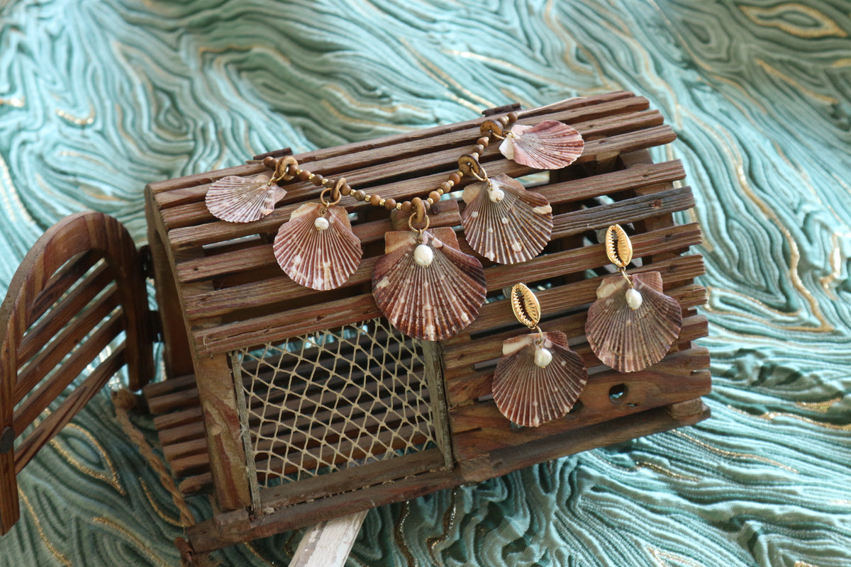 A handmade jewelry set featuring scallop shell charms and earrings displayed on a rustic wooden crate against a textured teal fabric background.